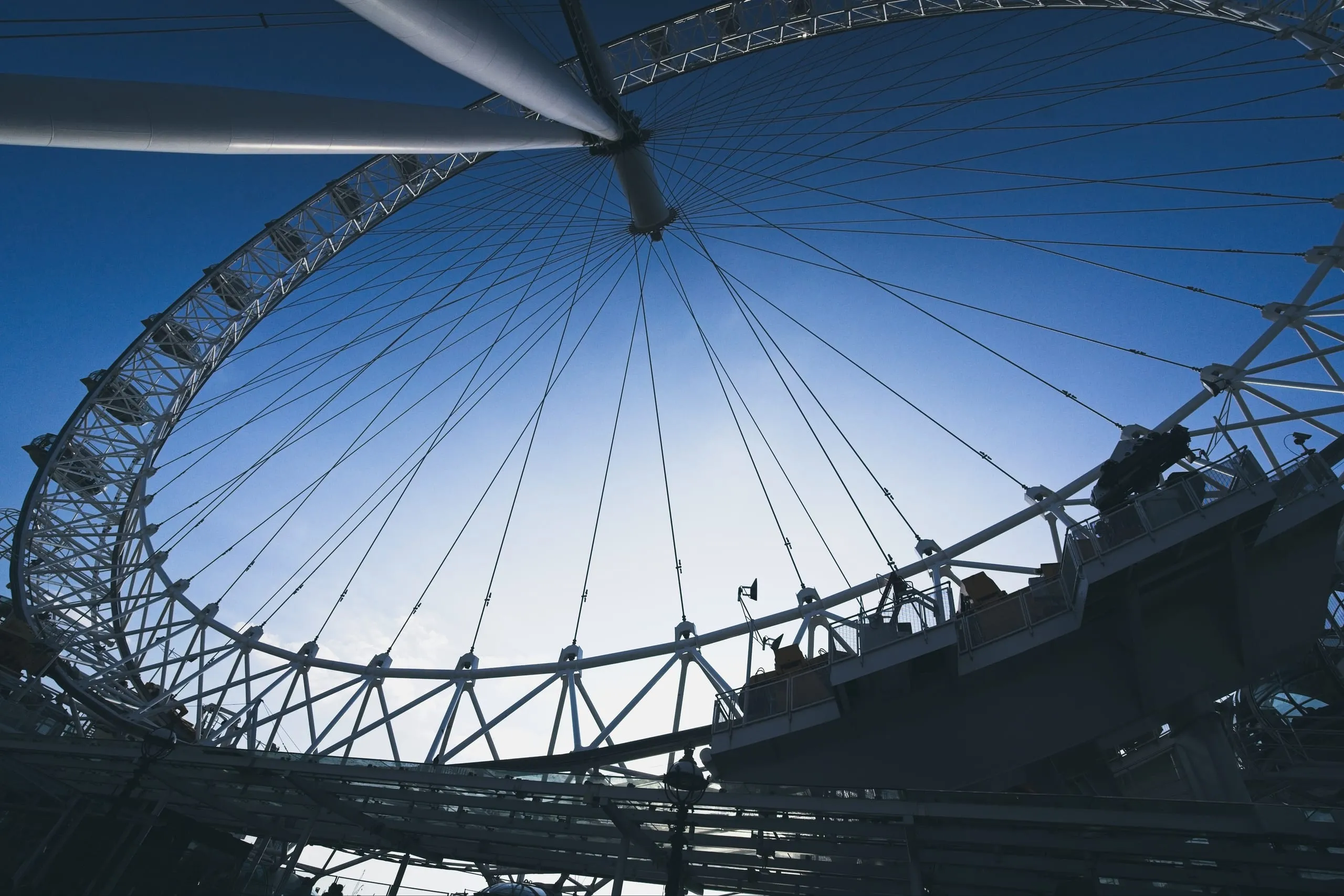 Photograph of the London Eye's rim and radiating support cables silhouetted against a clear sky, with one glass capsule visible at the edge, London, England.