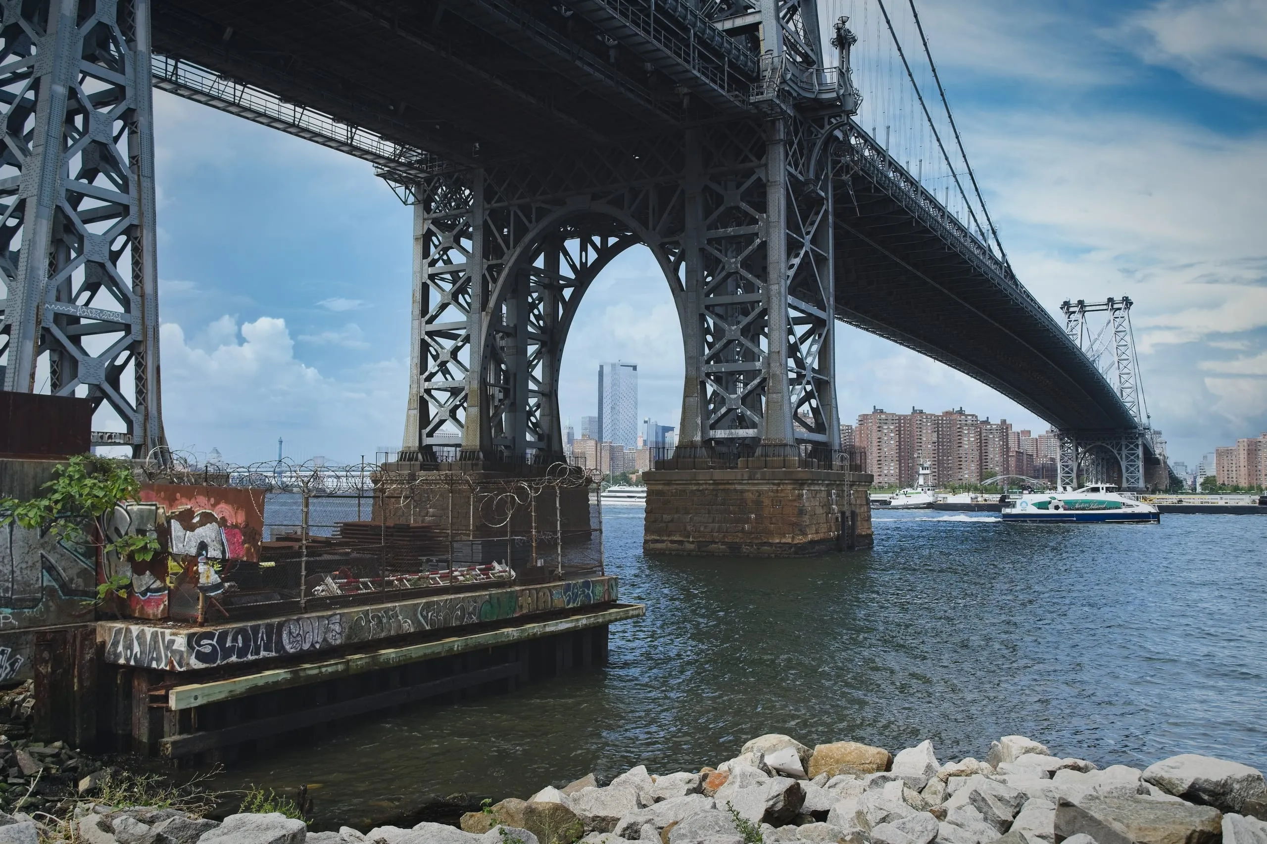 A color photograph of the steel underside and stone piers of the Williamsburg Bridge spanning the East River, with the Manhattan skyline and a passing NYC ferry in the background.